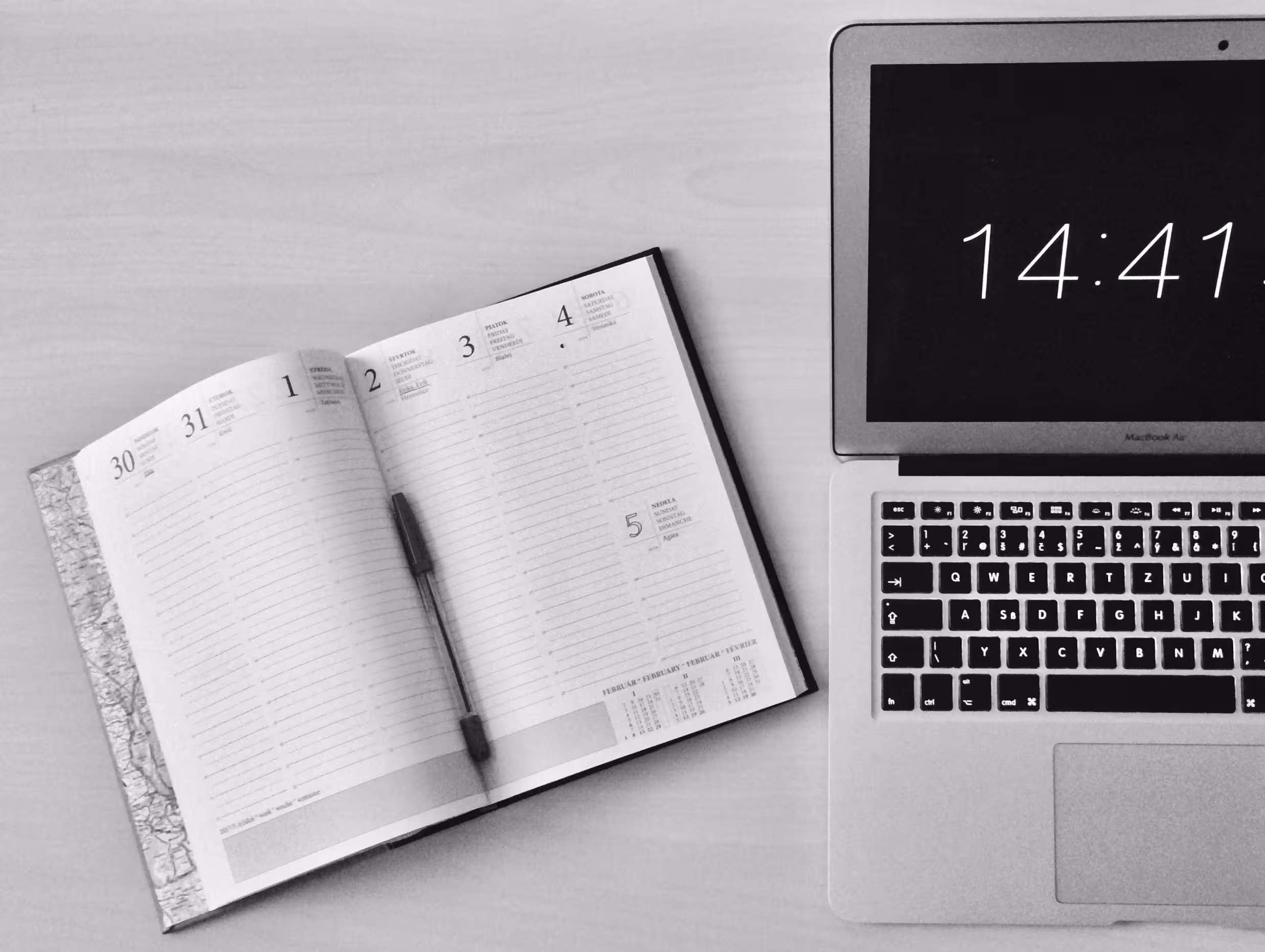 An open planner and pen beside a laptop displaying the time